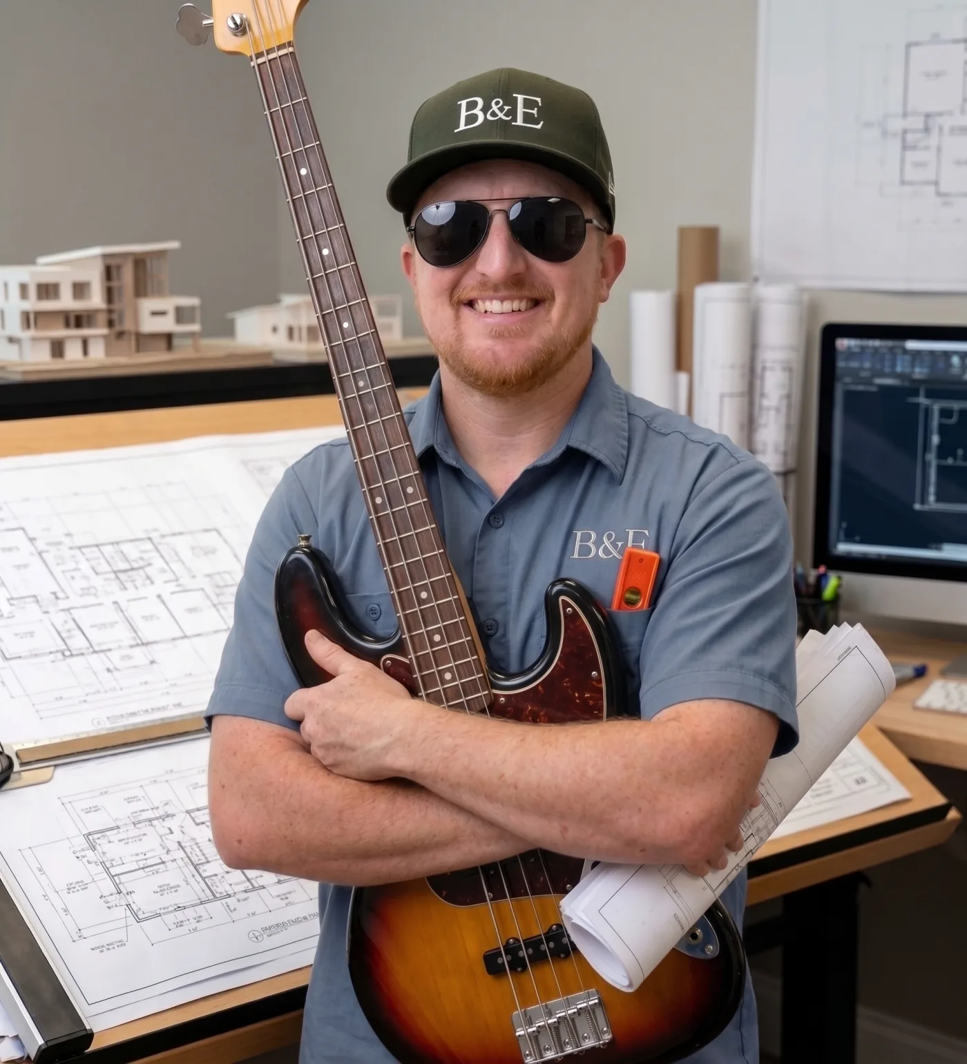 A B & D Elite Builders team member holding a bass guitar at the drafting table, project blueprints and a scale model behind.